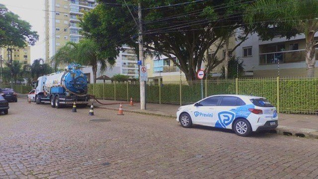 Caminhão e automóvel com marca Previni diante de edifício residencial em rua de paralelepípedos.