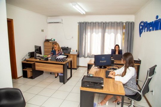 Pessoas em mesas de trabalho em escritório com marca Previni na parede.