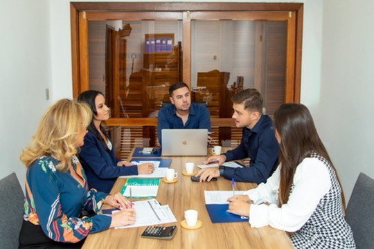 Cinco pessoas sentadas em torno de uma mesa, com papéis e notebook.